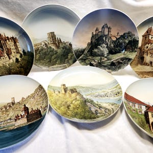 Può includere: Set di otto piatti in ceramica decorativi con illustrazioni panoramiche di castelli e paesaggi. Ogni piatto presenta un design unico con un castello o un edificio diverso, circondato da alberi, colline e fiumi. I piatti sono bianchi con una finitura lucida.