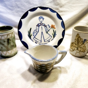 May include: A set of four ceramic mugs and a plate. The mugs have different designs, including a green and white geometric pattern, a brown and white leaf design, and a blue and white design with a girl holding a book. The plate has a blue and white border and a center design of a girl holding a book.