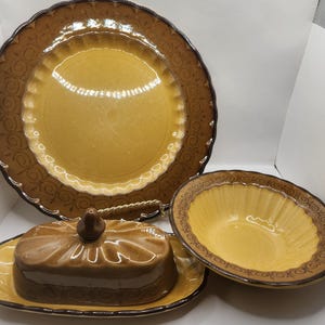 May include: A ceramic dinnerware set comprising a large plate, a bowl, and a covered butter dish. The set has a yellow centre with a brown decorative border. The butter dish has a matching lid with a small knob.