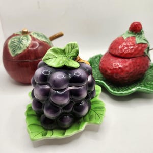 Peut inclure: Trois récipients en céramique en forme de fruits : une pomme rouge, une grappe de raisins violets et une fraise rouge. Chaque récipient a un couvercle et repose sur une base en forme de feuille verte. La fraise a une petite cuillère.