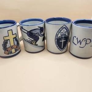 以下が含まれることがあります： 青と白のデザインが施されたセラミック製のマグカップが4つ。マグカップには、十字架、スキーヤー、十字架のある紋章、そして「LWPX」の文字が描かれています。