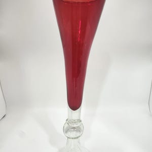 May include: A tall, elegant glass vase with a vibrant red upper section and a clear, decorative base. The vase has a flared top and a slender, elongated body, perfect for displaying flowers or as a decorative piece. The base features a bulbous design.