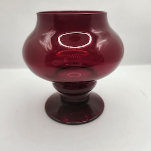 Peut inclure: Un bougeoir en verre rouge rubis avec un corps arrondi et une base en piédestal. Le verre a une finition brillante, reflétant la lumière. Le bougeoir est conçu pour contenir une seule bougie.