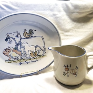 Puede incluir: Un plato de cerámica blanco con un borde azul presenta un diseño pintado a mano de animales de granja, incluyendo una vaca, un cerdo, un pollo y un pato. El plato tiene el texto "Free Depc. Sample" en letras azules. Una jarra de cerámica blanca con un borde azul y asa presenta un diseño pintado a mano de una vaca con manchas marrones.