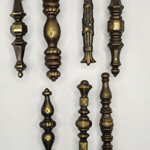 以下が含まれることがあります： さまざまな装飾デザインの、7つのアンティーク真鍮製引き出しプル。細長い形、球根状、円柱状など、さまざまな形状があり、詳細な装飾要素が特徴です。真鍮は暖かく、古びた風合いです。