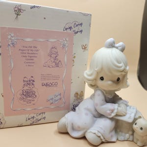 以下が含まれることがあります： 白い磁器製の女の子の置物。金色の髪に薄い紫色の白ドットのドレスを着ています。彼女は本を持っており、小さな白い子猫の隣に座っています。この置物はプレシャスモーメントのコレクションの一部であり、「You Fill the Pages of My Life」というタイトルが付けられています。