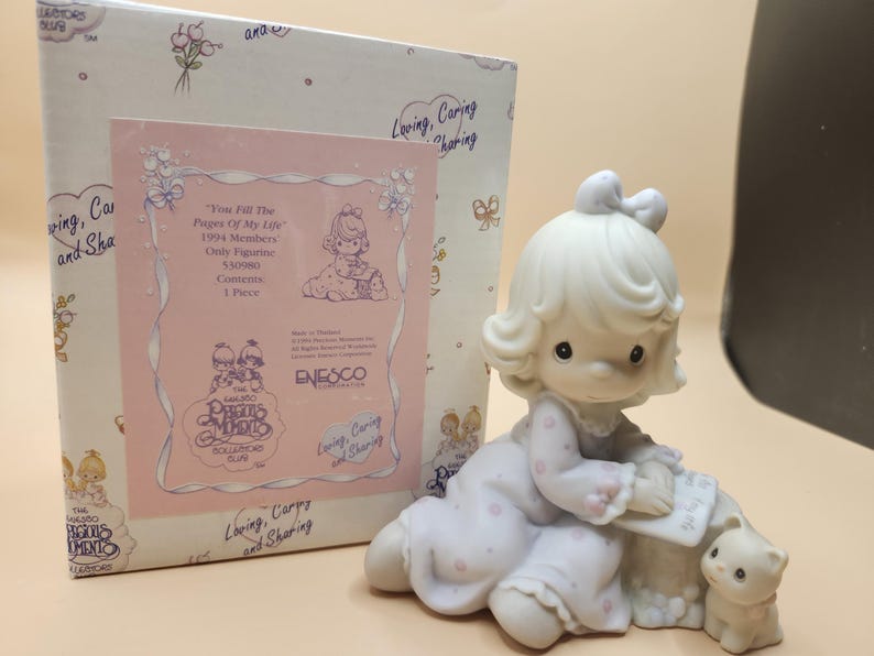 以下が含まれることがあります： 白い磁器製の女の子の置物。金色の髪に薄い紫色の白ドットのドレスを着ています。彼女は本を持っており、小さな白い子猫の隣に座っています。この置物はプレシャスモーメントのコレクションの一部であり、「You Fill the Pages of My Life」というタイトルが付けられています。