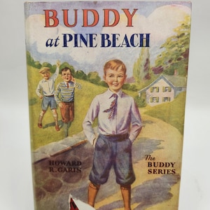 May include: A vintage book cover with the title "BUDDY at PINE BEACH" in blue and red lettering. The cover features an illustration of three boys in a rural setting with a house in the background. The book is part of "The BUDDY Series" by Howard R. Garis.