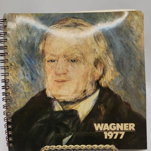 Puede incluir: Un libro encuadernado en espiral con una pintura de un hombre con cabello blanco y una chaqueta negra. El texto "WAGNER 1977" está impreso en la portada.