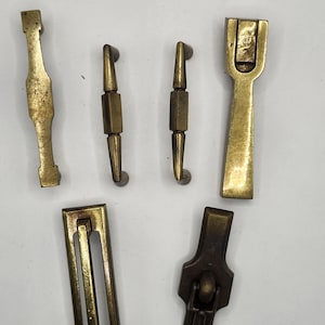 Könnte beinhalten: Eine Auswahl an antiken Messingbeschlägen, darunter Griffe und Verschlüsse. Die Teile variieren in Form und Größe und weisen ein angelaufenes, gealtertes Aussehen auf. Die Beschläge sind auf einem neutralen Hintergrund angeordnet, um die Details jedes Artikels hervorzuheben.