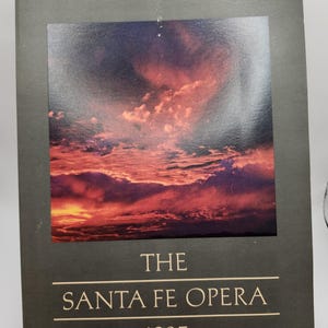 May include: A dark green book cover featuring a photograph of a sunset sky. The text "THE SANTA FE OPERA 1985" is printed in gold on the cover.