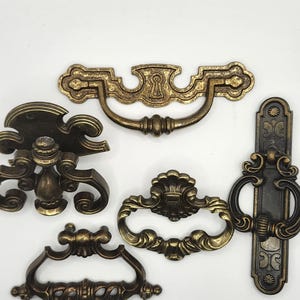 以下が含まれることがあります： アンティーク真鍮製の引き出し用つまみとハンドルの詰め合わせ。コレクションには、鍵穴型のプル、装飾的なディテールが施された丸いプル、装飾的なバックプレートが付いた垂直ハンドルなど、さまざまなスタイルが含まれています。ハードウェアはヴィンテージ感があります。