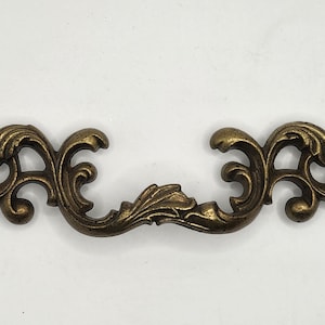 Puede incluir: Tirador de cajón antiguo de color bronce con un diseño curvo y ornamentado. La ferretería decorativa presenta detalles de hojas y vides. El tirador mide aproximadamente 15 cm de largo, perfecto para añadir un toque vintage a los muebles.