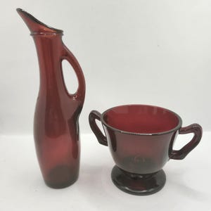 May include: A vintage ruby red glass pitcher with a curved handle and spout, and a matching sugar bowl with two handles. The pitcher has a slender, elegant design. The sugar bowl sits on a small pedestal base.