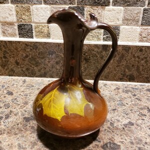 May include: A brown ceramic jug with a yellow leaf design. The jug has a round body and a long, thin neck.