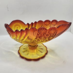 May include: An amber-colored, pressed glass pedestal bowl. The bowl has a scalloped rim and a starburst pattern on the sides. The base is a round, scalloped shape. The bowl is likely vintage and suitable for display or serving.
