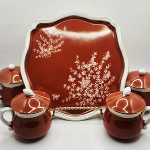 Pode incluir: Um conjunto de chá de cerâmica vermelha com uma bandeja e quatro xícaras com tampa. A bandeja e as xícaras apresentam um design de borboletas e flores brancas. A bandeja tem uma borda branca ondulada com acabamento dourado.