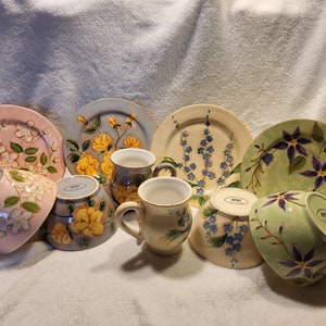 Peut inclure: Ensemble de vaisselle en céramique peinte à la main, comprenant des assiettes, des bols et des tasses. Les pièces présentent des motifs floraux de différentes couleurs, comme le rose, le jaune, le bleu et le violet, sur fond blanc. La vaisselle est disposée sur une surface blanche.