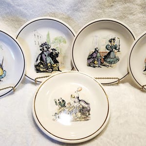 May include: Set of five white ceramic plates with gold trim. Each plate features a black and white illustration of people in various scenes.