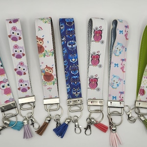 May include: Set of ten colorful keychains with owl designs. Each keychain features a different owl pattern on a white background. The keychains have silver metal rings and tassels.