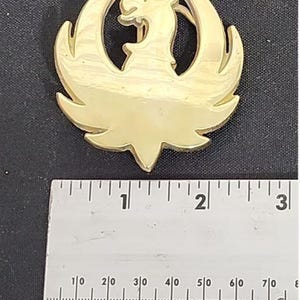May include: A gold-colored metal emblem featuring a stylized phoenix design. The phoenix is centered, with its wings spread and a detailed head. The emblem is approximately 2 inches wide, as measured by the ruler in the image.