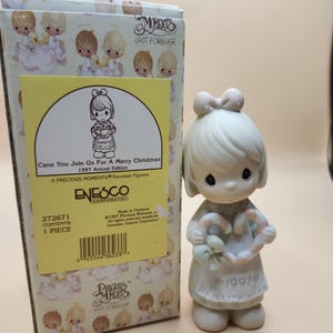 以下が含まれることがあります： 女の子がキャンディーケーンのハートを持っている、磁器製の置物です。彼女は緑色のサッシュと髪にリボンをつけた白いドレスを着ています。この置物はプレシャスモーメントのコレクションの一部であり、1997年製です。箱には「Cane You Join Us For A Merry Christmas 1997 Annual Edition」と書いてあります。