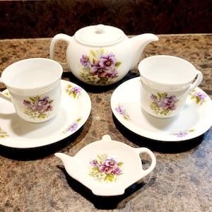 May include: A white tea set with a floral design. The set includes a teapot, two teacups, two saucers, and a small plate for the teapot.