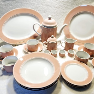 May include: A set of pink and white china dinnerware with a delicate gold trim. The set includes plates, bowls, cups, saucers, a teapot, a creamer, and a sugar bowl.