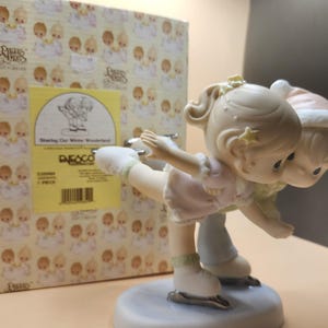 以下が含まれることがあります： プレシャスモーメンツのフィギュリン。2人の子供がスケートをしている。女の子はピンクのドレスと白いスケート靴を履いている。男の子は青と白の服を着て、茶色のスケート靴を履いている。フィギュリンは白い台座の上にあり、箱には「Sharing Our Winter Wonderland」と書かれている。