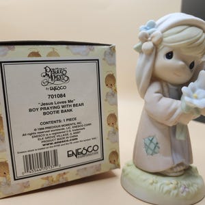 以下が含まれることがあります： 白いドレスを着た女の子が白い花を持っている、プレシャスモーメントの置物。置物は、クマの貯金箱と一緒に祈る男の子です。箱には「ジーザスラブズミー」と「701084」と書かれています。