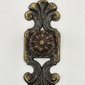 Puede incluir: Tirador de armario ornamentado de estilo antiguo con acabado en bronce y oro. El herraje decorativo presenta un pomo floral central y detalles de hojas en espiral. El tirador mide aproximadamente 13 cm de largo.
