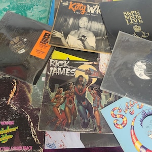 Może przedstawiać: Kolekcja płyt winylowych z widocznymi okładkami. Tytuły to m.in. "Kitty White", "Simple Minds Live", "Rick James" i "Rainbow Bridge" Jimiego Hendrixa. Płyty prezentują różne kolory i wzory, prezentując różne gatunki muzyczne.