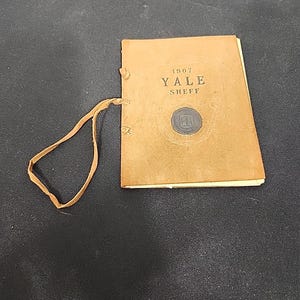 以下が含まれることがあります： 表紙に「1907 YALE SHEFF」と書かれた小さな茶色の小冊子。小冊子には革のストラップと円形のエンブレムが付いています。ページが見え、小冊子は暗い表面に置かれています。