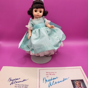 May include: A porcelain doll wearing a light blue dress with a white lace trim and a blue sash. The doll has black hair and blue eyes. The doll is standing on a white stand. The doll is accompanied by a card with the text "Welcome to the World of Madame Alexander" and a signature.