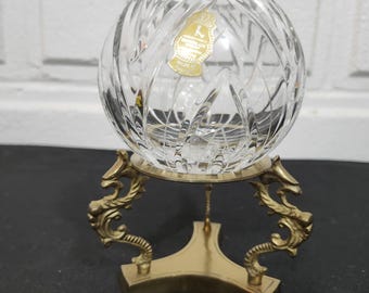 May include: A clear, cut-crystal bowl with a gold-colored sticker sits atop a decorative brass stand. The stand features ornate, curved arms and a square base. The bowl's intricate design catches the light, creating a sparkling effect.