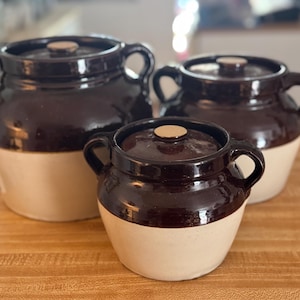 May include: A set of three two-toned ceramic crocks with lids. Each crock features a dark brown glazed upper section and a light cream-coloured lower section. The crocks have two side handles and a circular knob on the centre of each lid.