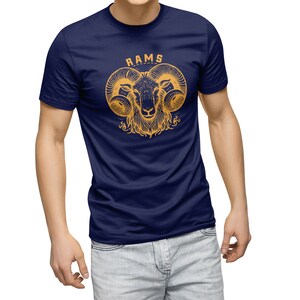 May include: Navy blue t-shirt featuring a gold illustration of a ram's head with the word "RAMS" above. The ram's head has large, curved horns and detailed facial features. The shirt is a classic crew neck style.