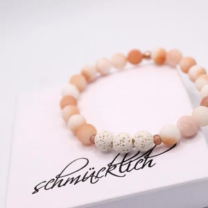 May include: A beaded bracelet with peach and white-colored stones. The bracelet is resting on a white box with the word "schmücklich" in a script font. The bracelet has three porous white beads.