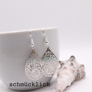 Peut inclure: Boucles d'oreilles argentées en forme de goutte d'eau avec des motifs de filigrane complexes, sur fond blanc. Les boucles d'oreilles sont présentées près d'une tasse en céramique blanche et d'un coquillage. Le mot "schmucklich" est imprimé en bas.