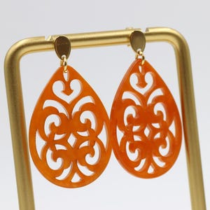 Könnte beinhalten: Ein Paar orangefarbene tropfenförmige Ohrringe mit einem filigranen Design. Die Ohrringe hängen an einem goldenen Ständer.