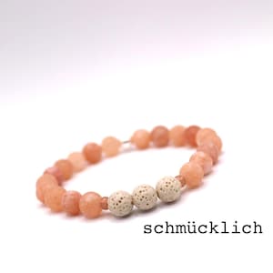 May include: A beaded bracelet featuring peach-colored beads and three textured, off-white beads. The bracelet is strung on a wire and is displayed against a white background. The word "schmücklich" is printed below the bracelet.