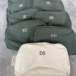 May include: A collection of small zippered pouches arranged in rows. Most pouches are sage green with white embroidered initials, while the front pouch is cream-colored with dark green embroidered initials DS. Each pouch features a top zipper.