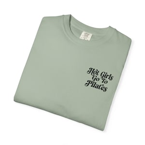 Puede incluir: Camiseta verde salvia con el texto negro "Hot Girls Go To Pilates". La camiseta está doblada y tiene cuello redondo. La etiqueta es visible. El texto está en una fuente elegante.