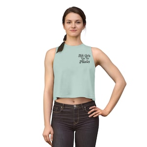 May include: A light green crop top with the text "Hot Girls Go To Pilates" in black script. The sleeveless top is paired with dark wash jeans. The woman is standing with her hand on her hip.
