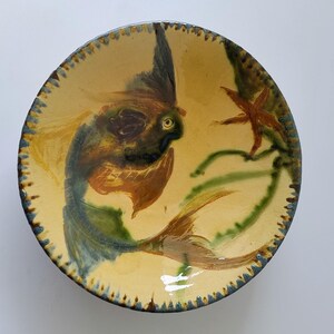 Puede incluir: Un cuenco de cerámica pintado a mano con un diseño de pez. El cuenco presenta un fondo amarillo con un pez en tonos azules, marrones y verdes. También se representan una estrella de mar y algas. El borde tiene un patrón azul y amarillo.