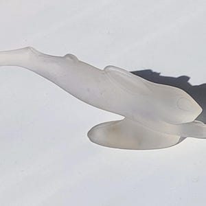 Könnte beinhalten: Eine Skulptur aus mattiertem Glas, die einen Vogel im Flug darstellt. Die Skulptur ist weiß und hat ein glattes, geschwungenes Design. Die Flügel des Vogels sind ausgebreitet und der Kopf ist nach oben geneigt. Die Skulptur ist auf einem abgerundeten Sockel montiert.