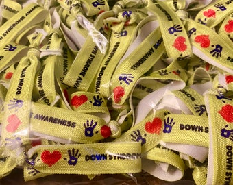 May include: A pile of yellow fabric hair ties with the words "DOWN SYNDROME AWARENESS" printed on them. Each tie features a red heart and a blue handprint. The ties are knotted and ready for use.