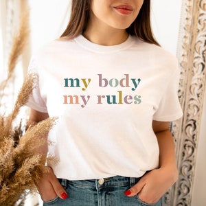 Puede incluir: Camiseta blanca con el texto "my body my rules" en letras de colores.