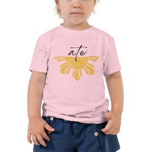 May include: A pink toddler t-shirt with a yellow sun graphic and the word "ate" in black lettering. The sun graphic is a stylized version of the Philippine sun.