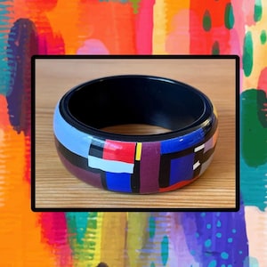 May include: A wide bangle bracelet featuring a smooth, glossy finish with a geometric pattern of bold, multi-colored blocks in shades of light blue, deep red, dark purple, black, and yellow, with a solid black interior lining.